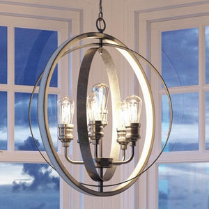 The UHP2211 Vintage Chandelier from Urban Ambiance’s Anchorage Collection, featuring a 30-3/4″ height and 28″ width with a galvanized steel finish, hangs in front of large windows overlooking a cloudy dusk sky, strikingly combining contemporary design with classic farmhouse charm.