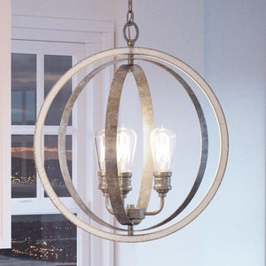 The UHP2213 Vintage Chandelier from the Anchorage Collection by Urban Ambiance, featuring a galvanized steel finish and measuring 24″H x 21-1/4″W, hangs near a window and bathes the room in a warm, vintage-inspired glow as the cityscape and sunset unfold outside.