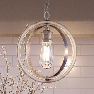 The UHP2215 Vintage Pendant from Urban Ambiance’s Anchorage Collection, featuring a 14-inch tall by 12-inch wide design in a galvanized steel finish, hangs elegantly with an exposed Edison bulb inside two intersecting metal rings; delicate flower branches and a white tile wall provide a charming backdrop.