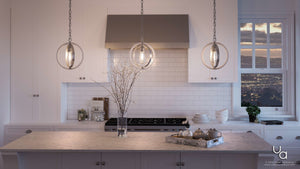 Modern white kitchen featuring a marble island illuminated by three vintage modern pendant lights, including the striking Urban Ambiance Anchorage Collection UHP2215 Vintage Pendant (14″H x 12″W) in a galvanized steel finish; also shown are a tray with teapot and cups, a vase with branches, open shelves displaying dishes, and a window offering a city view at sunset.