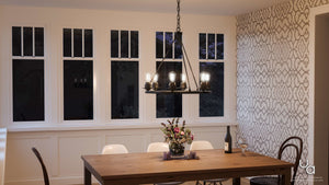 A wooden dining table adorned with a flower arrangement, candles, a wine bottle, and a glass sits beneath the Urban Ambiance UHP2243 Luxe Industrial Chandelier (27-1/2″H x 28″W, Charcoal Finish) from the Nashville Collection in a room with patterned wallpaper and large windows.