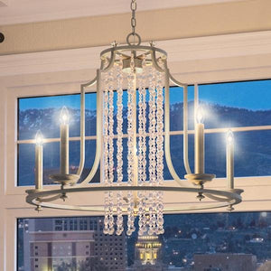The Urban Ambiance UHP2252 Mediterranean Chandelier from the Lexington Collection, measuring 24 inches high by 28 inches wide and finished in antique silver, features four candle-style lights and cascading crystal strands. It is suspended in front of a window that showcases a stunning cityscape with mountains silhouetted at dusk.