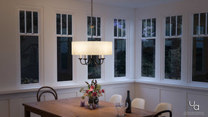 A dining room features a wooden table adorned with a flower centerpiece, wine bottle, and two glasses. Overhead, the Urban Ambiance Aurora Collection UHP2281 Transitional Chandelier (21-1/8″H x 21-5/8″W, Olde Bronze Finish) adds a striking modern farmhouse touch. The space is surrounded by tall windows revealing an evening view.