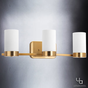 The UHP2293 Contemporary Bathroom Vanity Light from the Madison Collection by Urban Ambiance, featuring a brushed bronze finish and three frosted cylindrical glass shades, is mounted on a grey wall to provide a sophisticated accent that complements a crystal indoor chandelier for an elegant lighting ensemble.