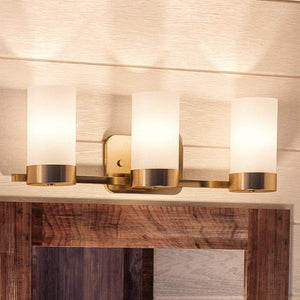 The Urban Ambiance UHP2293 Contemporary Bathroom Vanity Light from the Madison Collection, featuring a brushed bronze finish and three cylindrical frosted glass shades, is mounted above a doorway with wood trim, offering a subtle elegance reminiscent of an indoor crystal chandelier.