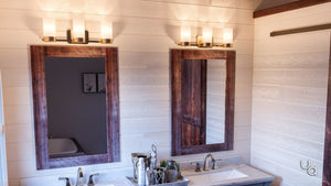 A double bathroom vanity features two wooden-framed mirrors, each illuminated by the Urban Ambiance UHP2293 Contemporary Bathroom Vanity Light from the Madison Collection, finished in brushed bronze and measuring 7.5” high by 24.75” wide, mounted above each mirror. Sinks are set below, with toiletries arranged between them, while an antique silver chandelier overhead casts a gentle glow throughout the space.