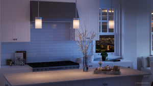 A modern kitchen features white cabinets, a marble island, backsplash tiles, and a night city view through the window. Above the island hang Urban Ambiance’s Madison Collection UHP2307 Transitional Pendant Lights (7″ x 5-1/8″) with etched white glass and a polished chrome finish, adding an elegant touch. A vase with branches and a tray with cups enhance the inviting atmosphere.