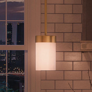 An Urban Ambiance UHP2309 Transitional Pendant Light from the Madison Collection, measuring 7″ x 5-1/8″ with a brushed bronze finish, hangs in front of beige subway tile near a window, with a cityscape at dusk visible outside.