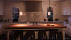 A modern kitchen features white cabinets, a subway tile backsplash, and a wooden island accompanied by three stools. On the island sit a cutting board with bread and a vase. Two windows are visible in the background, while above the island, stylish illumination is provided by Urban Ambiance’s Madison Collection UHP2309 Transitional Pendant Lights, each showcasing etched glass cylinder shades and finished in brushed bronze (7″ x 5-1/8″).