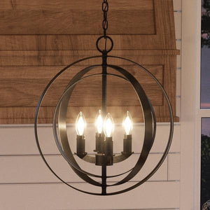 The UHP2324 Luxe Industrial Chandelier from Urban Ambiance’s Arezzo Collection, featuring an Olde Bronze finish and measuring 18-3/8″ high by 16″ wide, hangs in front of wood paneling and a white shiplap wall. Its black metal orb design with four candle-style bulbs evokes the elegance of an armillary sphere chandelier.