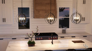 A modern kitchen featuring white cabinets, a marble island, a stove with distinctive red knobs, and two windows above the counter is illuminated by two round UHP2324 Luxe Industrial Chandeliers from Urban Ambiance’s Arezzo Collection in an Olde Bronze finish, adding armillary sphere-inspired elegance to the space.
