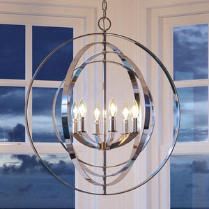 The Urban Ambiance UHP2328 Luxe Industrial Chandelier from the Arezzo Collection features interlocking rings in a polished nickel finish and six candle-style bulbs, elegantly displayed in front of expansive windows framing a cloudy evening sky.