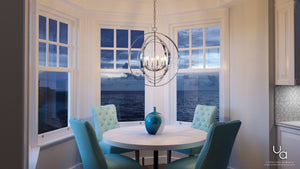 A round table with four turquoise chairs and a blue vase sits by bay windows overlooking the ocean at dusk, illuminated by the Urban Ambiance UHP2328 Luxe Industrial Chandelier from the Arezzo Collection, featuring interlocking rings in a polished nickel finish and measuring 30 inches high by 27-3/4 inches wide.