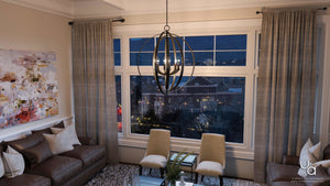 Modern living room with a brown sofa, abstract wall art, large windows offering a city view at dusk, grey curtains, and the Urban Ambiance UHP2330 Luxe Industrial Chandelier from the Arezzo Collection (30″H x 27-3/4″W) in an elegant Olde Bronze finish.