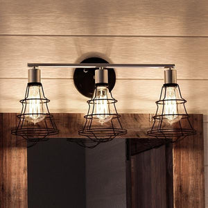 Three exposed bulbs with wire cage shades, mounted on the Urban Ambiance UHP2421 Vintage Bathroom Vanity Light in a charcoal finish from the Syracuse Collection, are installed horizontally above a mirror; a wooden frame and horizontal wall panels can be seen in the background.
