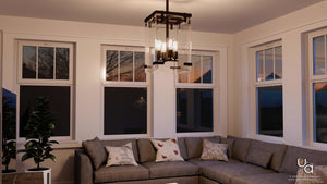 A modern living room featuring a gray sectional sofa with assorted pillows, a potted plant, expansive windows framing the sunset, and the Urban Ambiance UHP2442 Modern Farmhouse Modern Chandelier from the Bristol Collection—a contemporary cube ceiling light fixture in an Olde Bronze finish (28-3/8″H x 14-3/4″W SQ).