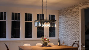 A modern dining room featuring a wooden table, four white chairs, a floral centerpiece, a bottle of wine with glasses, and a geometric-patterned accent wall, all illuminated by the Urban Ambiance UHP2444 Modern Farmhouse Square Chandelier from the Bristol Collection in an Olde Bronze finish.