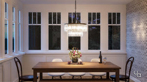 A wooden dining table with four white chairs is set with flowers, candles, and a wine bottle beneath the Urban Ambiance UHP2462 Traditional Chandelier from the Lille Collection, featuring a prismatic glass design and Olde Bronze finish, in a room with large windows and patterned wallpaper.