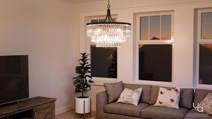 A living room featuring a gray sofa, potted plant, TV, and the Urban Ambiance UHP2464 Traditional Chandelier from the Lille Collection (29-1/4″ H x 28″ W, Olde Bronze Finish). Three windows reveal an evening sky.