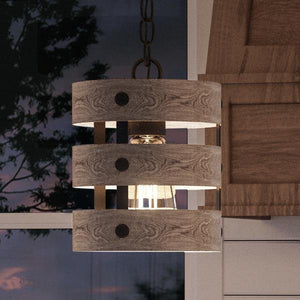 The UHP2472 Modern Farmhouse Pendant from Urban Ambiance’s Adelaide Collection brings a rustic modern touch indoors, featuring a wooden-inspired design with three circular bands in a charcoal finish. Suspended near a window, its open cage aesthetic complements views of surrounding trees and an adjacent wooden cabinet, amplifying the natural ambiance of the space.
