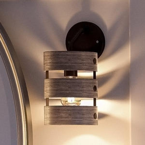 The UHP2480 Modern Farmhouse Bath / Wall Light from Urban Ambiance’s Adelaide Collection features a charcoal finish and measures 12 inches high by 6 inches wide. Showcasing a cylindrical, wood-textured shade with horizontal slats, this wall-mounted fixture casts distinctive shadows to create an inviting, farmhouse-inspired ambiance that blends natural warmth with contemporary style—perfect for rustic bathrooms.