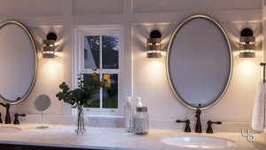 A double bathroom vanity features two oval mirrors with a window centered between them, adorned by Urban Ambiance’s UHP2480 Modern Farmhouse Bath/Wall Light from the Adelaide Collection in a charcoal finish. The marble countertop includes two sinks and is styled with decorative accents like a vase of greenery and glass jars.