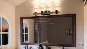 A bathroom vanity with a spacious wooden-framed mirror is illuminated by the UHP2486 Modern Farmhouse Bath Fixture from Urban Ambiance’s Adelaide Collection, featuring a sleek charcoal finish above, while arched windows and an array of toiletries on the sink countertop sit beneath a stylish rustic ceiling fan that adds both charm and comfort to the space.