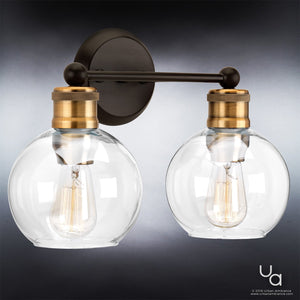 The Urban Ambiance UHP2500 Vintage Industrial Bath Fixture from the Belfast Collection features two clear globe glass shades with exposed filament bulbs and an Olde Bronze finish accented by brass metal details, creating a striking look against a neutral background. Measuring 10-7/8″ in height and 15-1/2″ in width, this wall-mounted fixture combines vintage industrial style with modern elegance.