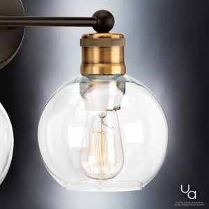 The UHP2500 Vintage Industrial Bath Fixture from the Belfast Collection by Urban Ambiance, featuring clear globe glass and an exposed filament bulb, is showcased with an Olde Bronze finish arm mounted on a wall against a gradient gray background. Measuring 10-7/8″ high by 15-1/2″ wide.
