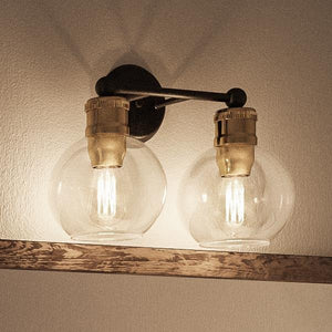 The Urban Ambiance UHP2500 Vintage Industrial Bath Fixture from the Belfast Collection features an elegant Olde Bronze finish and two exposed filament bulbs set within clear globe glass shades, beautifully mounted above a wooden shelf.
