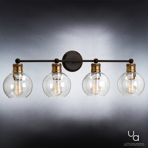 The Urban Ambiance UHP2504 Vintage Industrial Bath Fixture from the Belfast Collection features four clear glass globe shades, exposed bulbs, and striking metal accents in an Olde Bronze finish. This wall-mounted light has a vintage-inspired aesthetic and measures 10-7/8″ high by 33-1/2″ wide, shown photographed against a gradient gray background.