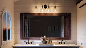 Double-sink bathroom vanity with a large wooden-framed mirror, featuring the Urban Ambiance UHP2504 Vintage Industrial Bath Fixture from the Belfast Collection in an Olde Bronze finish mounted above, and various toiletries arranged on the counter.