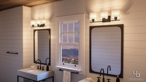 A modern bathroom features double sinks, rectangular mirrors, and the Urban Ambiance UHP2513 Cosmopolitan Bathroom Vanity Light from the Tacoma Collection. This wall-mounted fixture showcases an elegant Olde Bronze finish with Mylar cylinder shades. The space is illuminated by a central window offering views of the city and mountains at dusk.