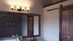 A bathroom vanity with a wooden-framed mirror is paired with the Urban Ambiance UHP2515 Cosmopolitan Bathroom Vanity Light from the Tacoma Collection, featuring Mylar cylinder shades in an Olde Bronze finish. A sliding barn door completes the look, while soap, a toothbrush holder, and a potted plant rest on the countertop. Light wood walls surround the space.