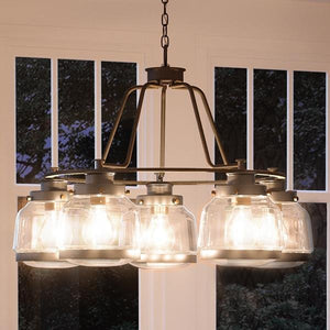 A striking Urban Ambiance UHP2531 Luxe Industrial Chandelier from the Nottingham Collection, featuring an Olde Bronze finish and six exposed bulbs cradled in glass shades, hangs from a chain in front of large windows.