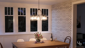 A dining room featuring a wooden table and three chairs is illuminated by the Urban Ambiance UHP2531 Luxe Industrial Chandelier from the Nottingham Collection, measuring 19.25 inches high by 26 inches wide, in an Olde Bronze finish. The scene is completed with a flower vase, a wine bottle, and a glass on the table, set against patterned wallpaper and large windows at dusk.