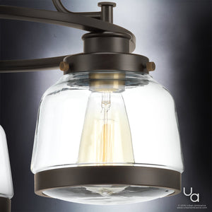 Close-up of the Urban Ambiance UHP2533 Luxe Industrial Chandelier from the Nottingham Collection, showcasing its Olde Bronze finish, clear artisan-style glass globe, exposed bulb, and dark metal accents against a gray background; measures 18″ high by 22″ wide.