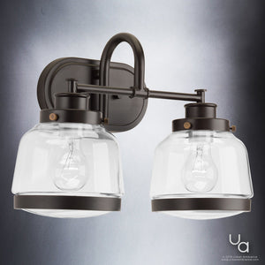 The UHP2537 Luxe Industrial Bathroom Vanity Light by Urban Ambiance features two clear glass shades and a vintage-inspired bronze metal fixture in an Olde Bronze finish. Measuring 11.25″ high by 16.25″ wide, this distinctive piece from the Nottingham Collection is mounted on a rectangular backplate against a gradient gray background. Perfect as a schoolhouse bath light, its timeless metal fittings lend classic charm to any space.