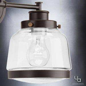 Close-up of the Urban Ambiance UHP2537 Luxe Industrial Bathroom Vanity Light from the Nottingham Collection, featuring an Olde Bronze finish, clear glass shade, exposed bulb, and vintage metal fittings against a gray background.