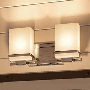 The Urban Ambiance UHP2601 Transitional Bathroom Vanity Light from the Modena Collection features a polished chrome finish and two etched opal glass cube shades that emit warm light, creating a modern accent against a beige wall.