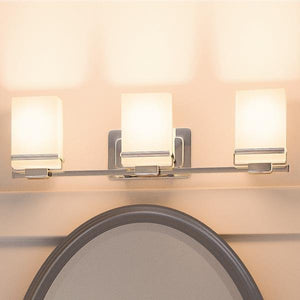 The Urban Ambiance UHP2603 Transitional Bathroom Vanity Light from the Modena Collection, featuring three opal glass cylinder shades and a sleek polished chrome finish, is mounted above an oval mirror on a beige wall, serving as a stylish centerpiece for the bathroom.