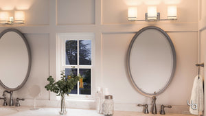 Modern bathroom with double sinks, oval mirrors, stylish wall lights, a small window, and a countertop adorned with plants, jars, and towels. Elegant bathroom lighting from Urban Ambiance’s Modena Collection features the UHP2603 Transitional Bathroom Vanity Light—a 6.5-inch-high by 22.125-inch-wide fixture in polished chrome—adding a bright and welcoming atmosphere with its opal glass cylinder design.