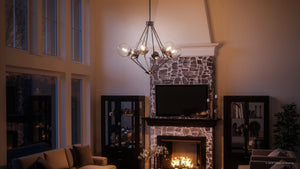 Experience a modern living room with high ceilings, a stone fireplace, and a TV above the mantle, accented by glass cabinets on either side. Overhead, the Urban Ambiance UHP2624 Modern Chandelier from the Costa Mesa Collection—a striking 24-7/8″ x 34″ fixture—features clear glass globes and a polished chrome finish for an elegant industrial touch.