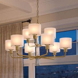 A UHP2630 XL Cosmopolitan Chandelier from the Oxford Collection by Urban Ambiance, featuring a Palladian Gold finish with ten cylindrical white lampshades and subtle marble accents, hangs from the ceiling in a room with large windows and beige curtains.