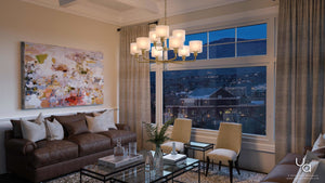 Modern living room featuring a brown sofa, abstract wall art, glass coffee table with marble accents, two beige chairs, and the UHP2630 XL Cosmopolitan Chandelier from Urban Ambiance’s Oxford Collection (25″H x 36″W, Palladian Gold Finish) suspended overhead. A large window frames a stunning dusk view of the cityscape and surrounding mountains.