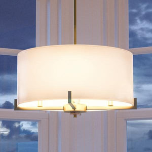 A sleek UHP2632 Cosmopolitan Drum Chandelier by Urban Ambiance, featuring a Palladian Gold finish from the Oxford Collection, hangs elegantly in front of expansive windows at dusk.