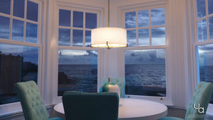 A round white table with marble accents and teal chairs sits by large windows overlooking the ocean at dusk, illuminated by the Urban Ambiance UHP2632 Cosmopolitan Drum Chandelier in a Palladian Gold finish from the Oxford Collection, suspended above.