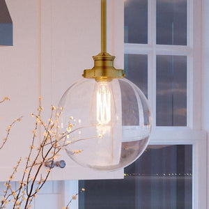The Urban Ambiance UHP2643 Vintage Pendant from the Glasgow Collection, featuring a clear glass globe and exposed bulb with a native brass finish (12-3/4″H x 9-3/4″W), hangs in front of a white window and cabinet beside decorative branches, bringing a touch of vintage modern lighting to the space.