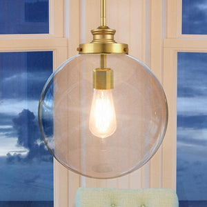 The UHP2645 Vintage Pendant from the Glasgow Collection by Urban Ambiance, featuring a 15-inch high by 12-inch wide round glass globe and a native brass finish, hangs illuminated in front of expansive windows revealing a cloudy dusk sky, lending an elegant modern accent to the space.