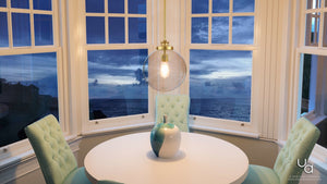 A round white table with a vase sits by bay windows overlooking the ocean at sunset, surrounded by four tufted chairs and softly illuminated by the Urban Ambiance UHP2645 Vintage Pendant from the Glasgow Collection, featuring a 15″ height, 12″ width, and native brass finish—adding a touch of modern lighting to the serene setting.
